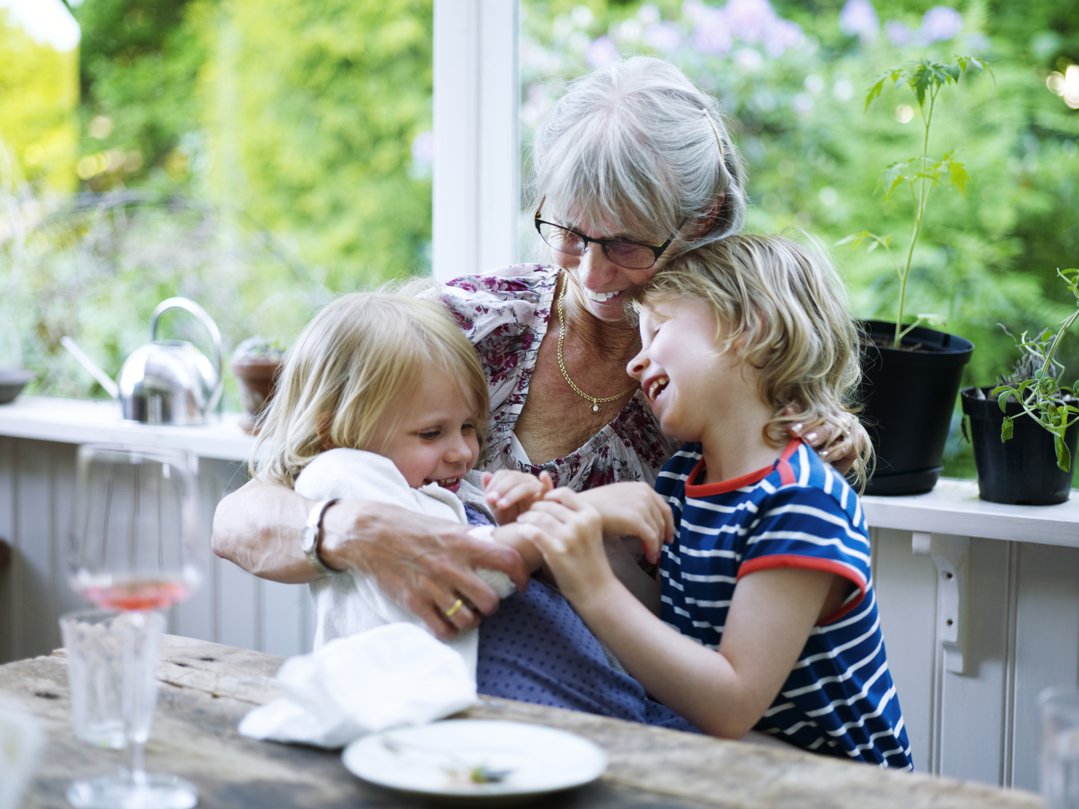 En leende äldre kvinna sitter med två små skrattande barn i knäet vid ett matbord.