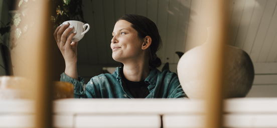 Ung kvinna tittar ut genom fönster, håller kaffekopp i handen