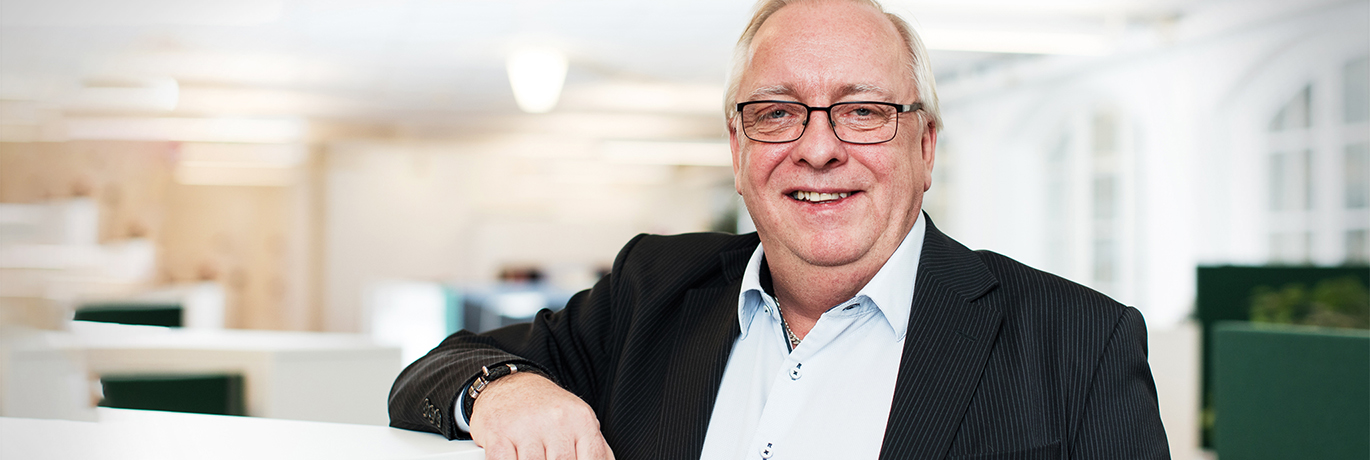 En leende äldre man i kontorsmiljö står och lutar sig mot ett skrivbord.